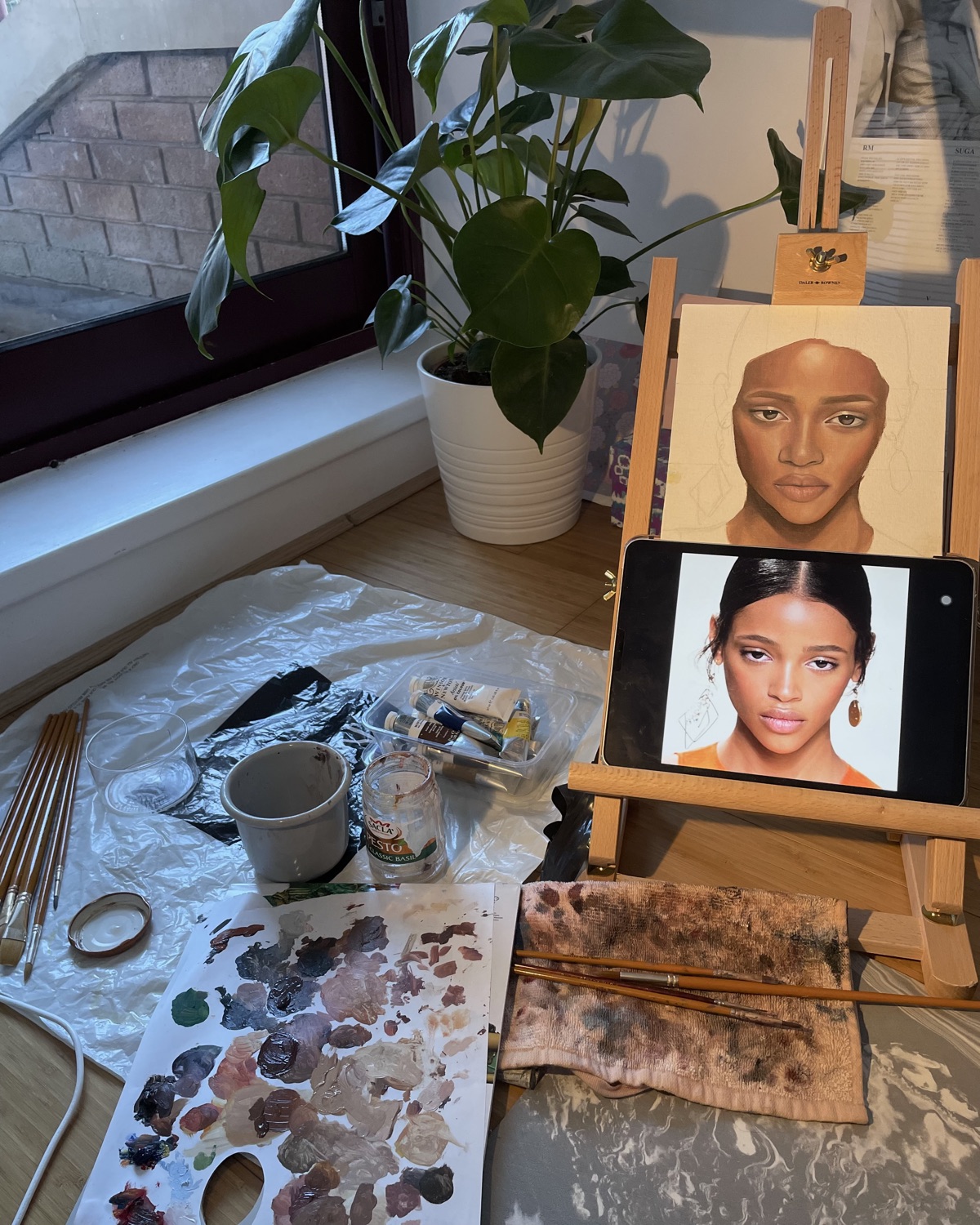 An easel with a portrait in progress beside a window and a monstera plant